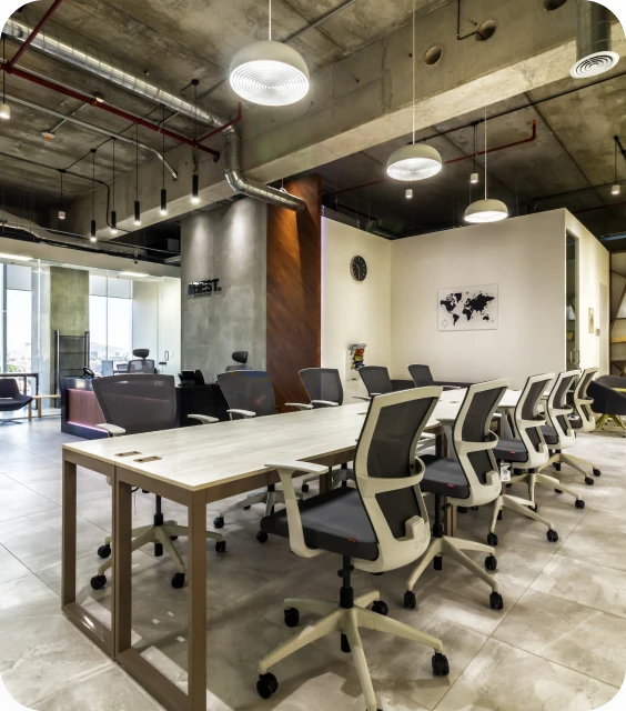 Sala de juntas alargada con mesa de madera clara y sillas grises ergonómicas, techo industrial con iluminación colgante