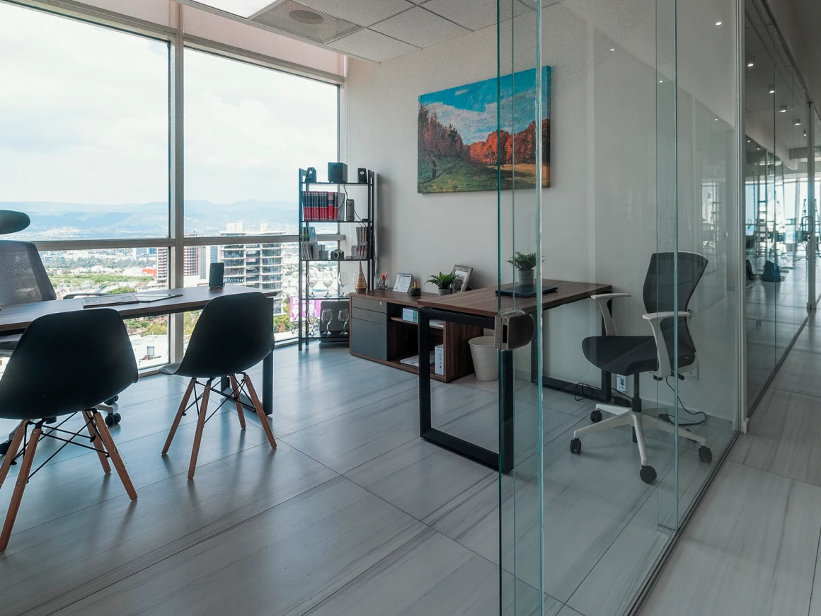 Oficina privada con escritorio de madera, sillas Eames negras, librero y cuadro de paisaje en la pared, con vista a la ciudad