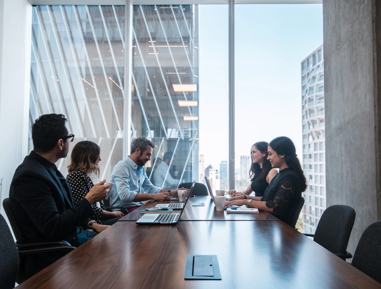 Equipo de trabajo reunido en una sala de juntas con vista a la ciudad, usando laptops sobre una mesa de madera